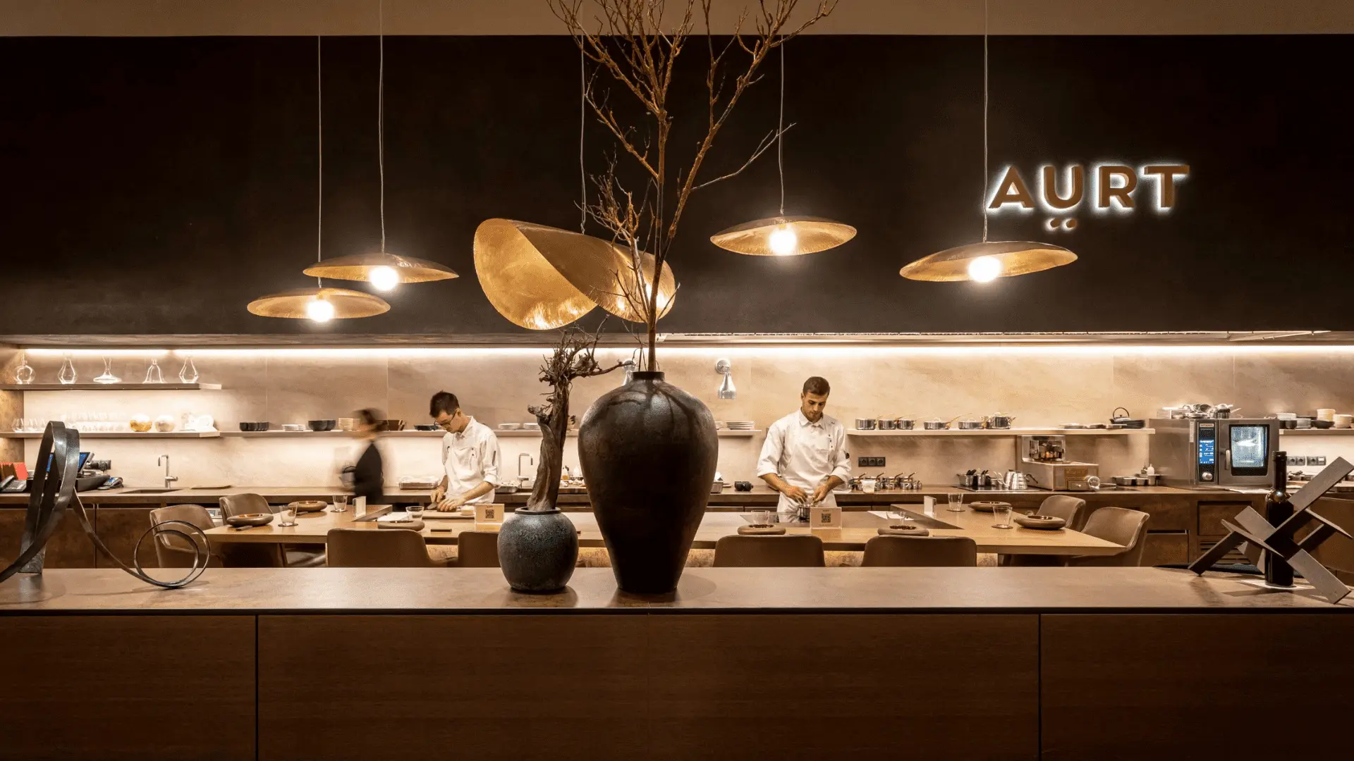 Cocina abierta de un restaurante con mesas alrededor. En primer plano un mueble con dos jarrones y troncos secos. cinco lámparas cuelgan del techo y en la pared oscura el letrero de AURT iluminado