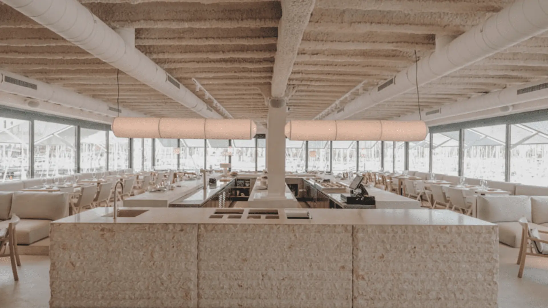 Interior de un restaurante con la barra de piedra en el centro y a los lados las mesas con sillones. Todo en tonos blanco roto