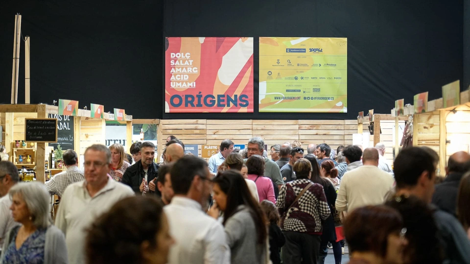 Personas en en la feria orígens de barcelona, cartel con el nombre orígens y dolç, salat, amarg, àcid y umami