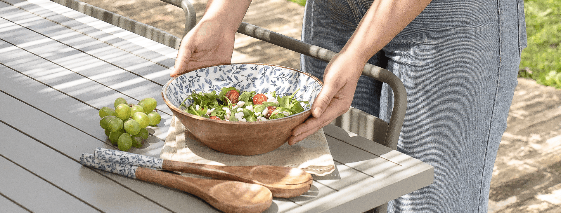 Manos sosteniendo una ensaladera al aire libre sobre una mesa con cucharas de madera y uvas verdes